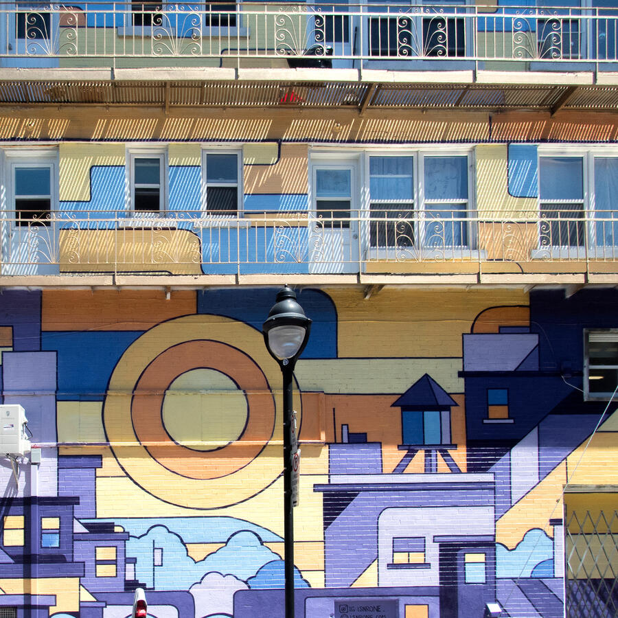 Détail de la murale peinte par Lucas Saenger sur le bâtiment. On y voit des bâtiments devant un soleil couchant.